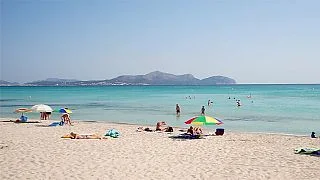 Mediterranes Natursteinhaus mit Terrasse, blauem Sonnenschirm, Holzmöbeln, Liegesessel und blühenden Pflanzen, umgeben von Zypressen und Palmen unter blauem Himmel