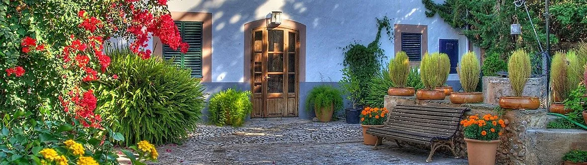 Mediterraner Innenhofgarten mit blauer Wand, Holztür, Steinbank, Topfpflanzen mit orangen und gelben Blumen und leuchtend roter Bougainvillea, die links hinaufklettert