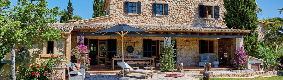 Mediterranes Natursteinhaus mit Terrasse, blauem Sonnenschirm, Holzmöbeln, Liegesessel und blühenden Pflanzen, umgeben von Zypressen und Palmen unter blauem Himmel